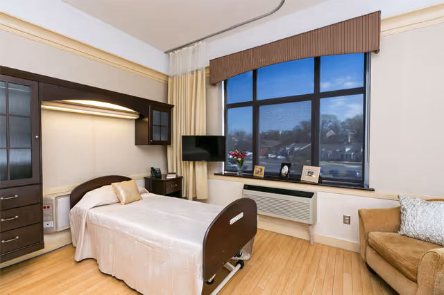 A nursing facility room with a single hospital bed covered in a beige bedspread, a wooden cabinet with glass doors above the bed, a small bedside table with a phone, a flat-screen TV mounted near a large window with beige curtains and a brown valance, a vase with flowers and framed photos on the windowsill, a beige armchair with a decorative pillow, and light wood flooring.