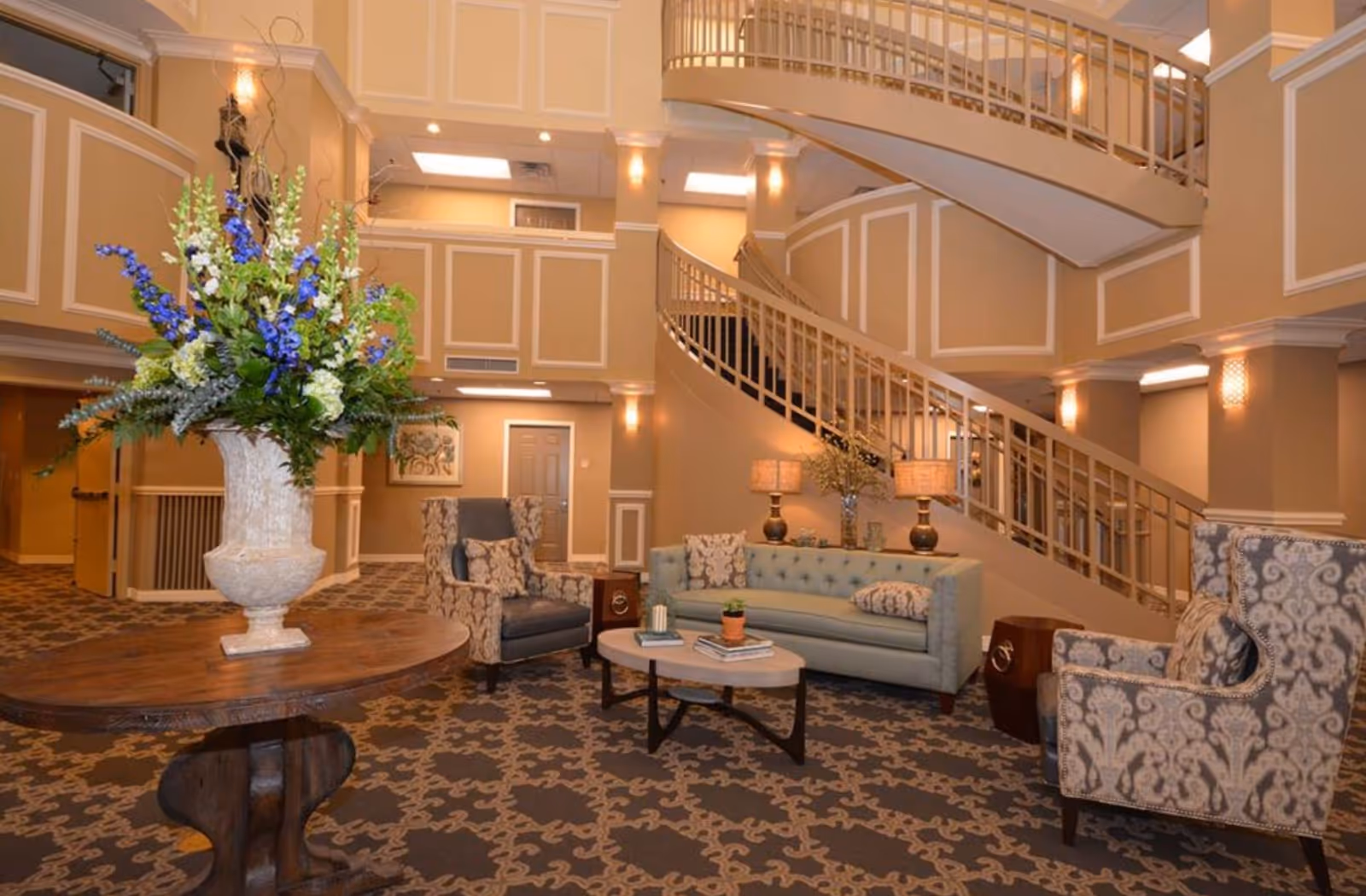 A spacious and elegantly decorated common area in a retirement community featuring a large floral arrangement on a wooden round table, patterned carpet, a light blue sofa with cushions, two patterned armchairs, a coffee table with books and a small plant, two table lamps, and a curved staircase with railings leading to an upper floor.