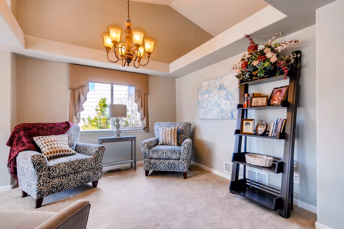 A cozy living room area with two patterned armchairs, one draped with a red throw blanket and both with decorative pillows. Between the chairs is a small table with a lamp in front of a window with beige curtains. On the right side, there is a black leaning bookshelf decorated with framed photos, books, and a basket, topped with a floral arrangement. A chandelier with warm lights hangs from the ceiling.