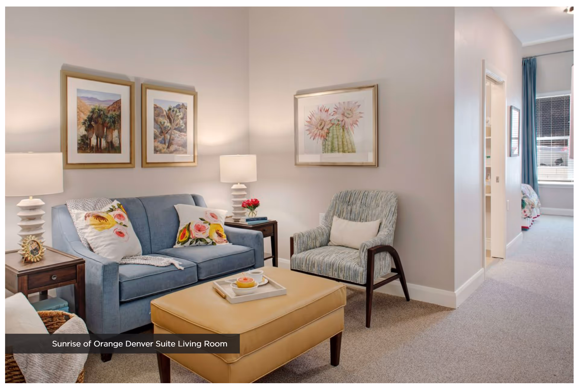 A cozy living room with a blue sofa adorned with floral pillows, a patterned armchair with a white cushion, a tan ottoman with a tray holding a cup and a pastry, two side tables with white lamps, and framed botanical artwork on the walls. A hallway leads to a bedroom with a window and blue curtains.