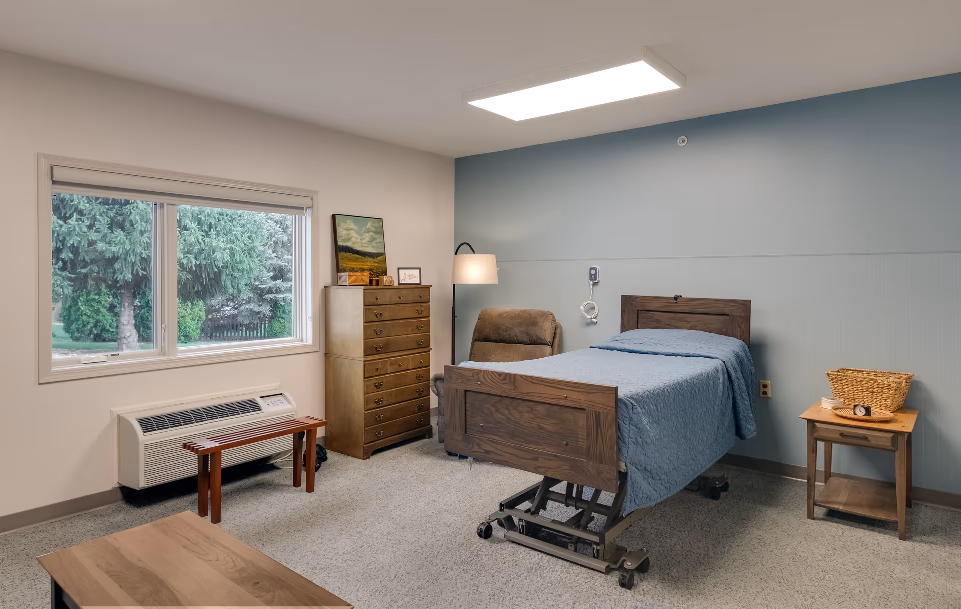 A senior living facility bedroom with a hospital-style bed covered in a blue quilt, a brown recliner chair, a wooden chest of drawers with framed pictures and a painting on top, a small wooden side table with a basket and a clock, a wooden bench under a large window showing green trees outside, and a wall-mounted air conditioning unit below the window.