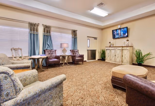 A cozy living room area in a senior care facility with patterned carpet, upholstered armchairs, a wooden side table with a lamp, a large ottoman, and a cabinet with a flat-screen TV mounted above it. The room has beige walls, large windows with blue curtains, and potted plants near the cabinet.