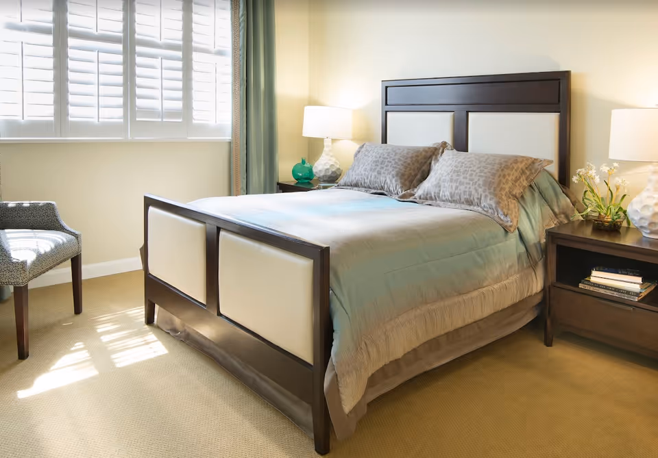 A bright bedroom featuring a neatly made bed with a dark wooden frame and light-colored padded panels. The bed has two patterned pillows and a light blue and beige comforter. On either side of the bed are dark wooden nightstands with white lamps, a small green vase, and a flower arrangement. A patterned chair is positioned near a window with white shutters and green curtains, allowing sunlight to stream into the room.