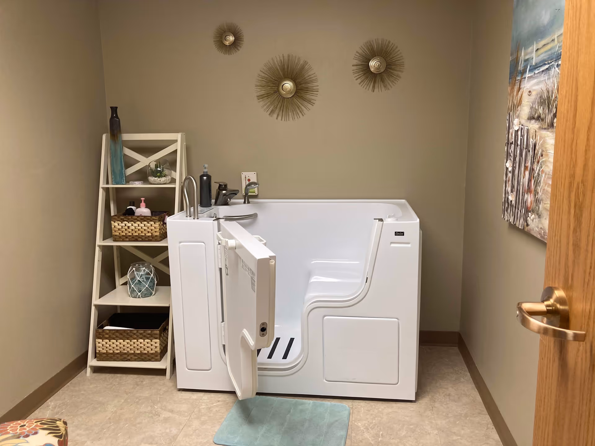 A small bathroom with a white walk-in bathtub featuring an open door. To the left of the tub is a white shelving unit holding decorative items and toiletries. The walls are beige with three decorative sunburst wall hangings above the tub and a beach-themed painting on the right wall. A light blue bath mat is placed in front of the tub.