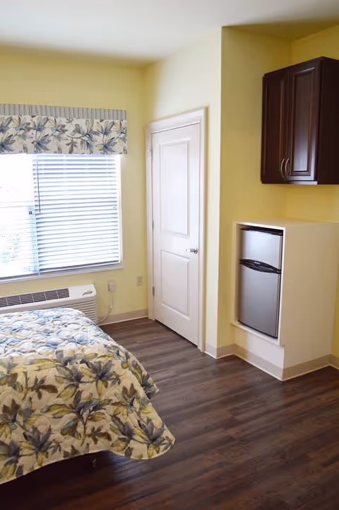 A bright bedroom with a bed covered in a floral patterned quilt, a window with blinds and a floral valance, a white door, a small refrigerator built into a white cabinet, and dark wood flooring.