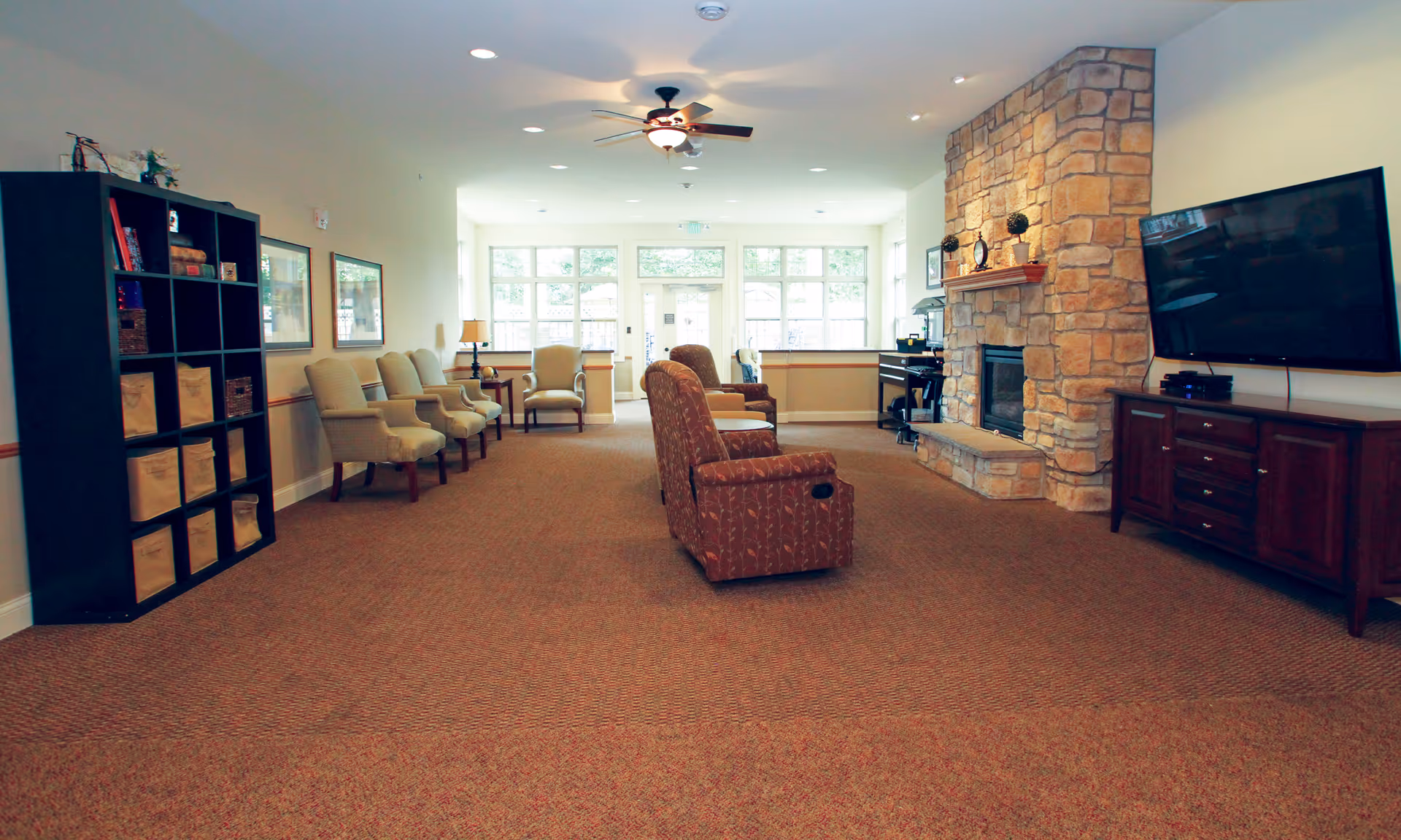 Spacious senior living common room with chairs and armchairs facing a stone fireplace and wall-mounted TV.