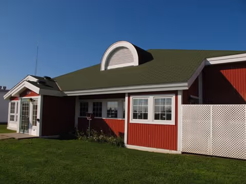 Exterior view of a single-story red building with white trim and a green roof under a clear blue sky. The building has multiple windows and a white lattice fence on the right side, with a well-maintained green lawn in front.