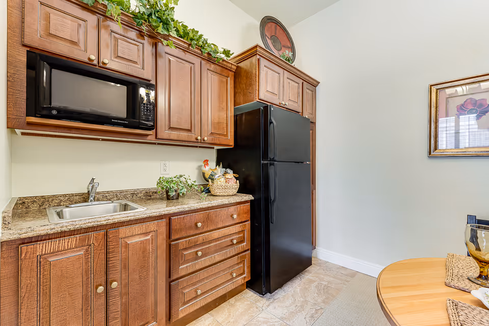 A small kitchen area with wooden cabinets, a black microwave mounted above the countertop, a stainless steel sink, and a black refrigerator. There are decorative plants and a basket on the countertop. Part of a round wooden dining table with placemats and a glass is visible on the right side. The walls are light-colored, and there is a framed floral picture hanging on the wall.