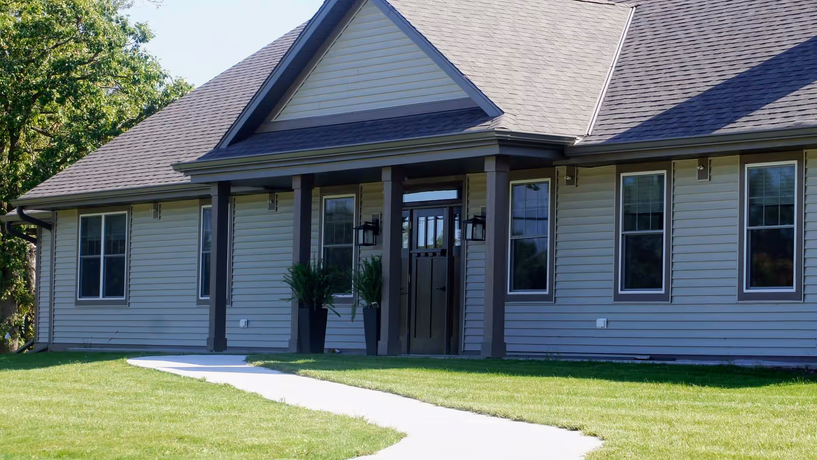 Exterior view of a single-story building with beige siding, a dark shingled roof, and a covered entrance supported by columns. There are several windows and two potted plants flanking the front door. A curved concrete walkway leads up to the entrance, and green grass surrounds the building.
