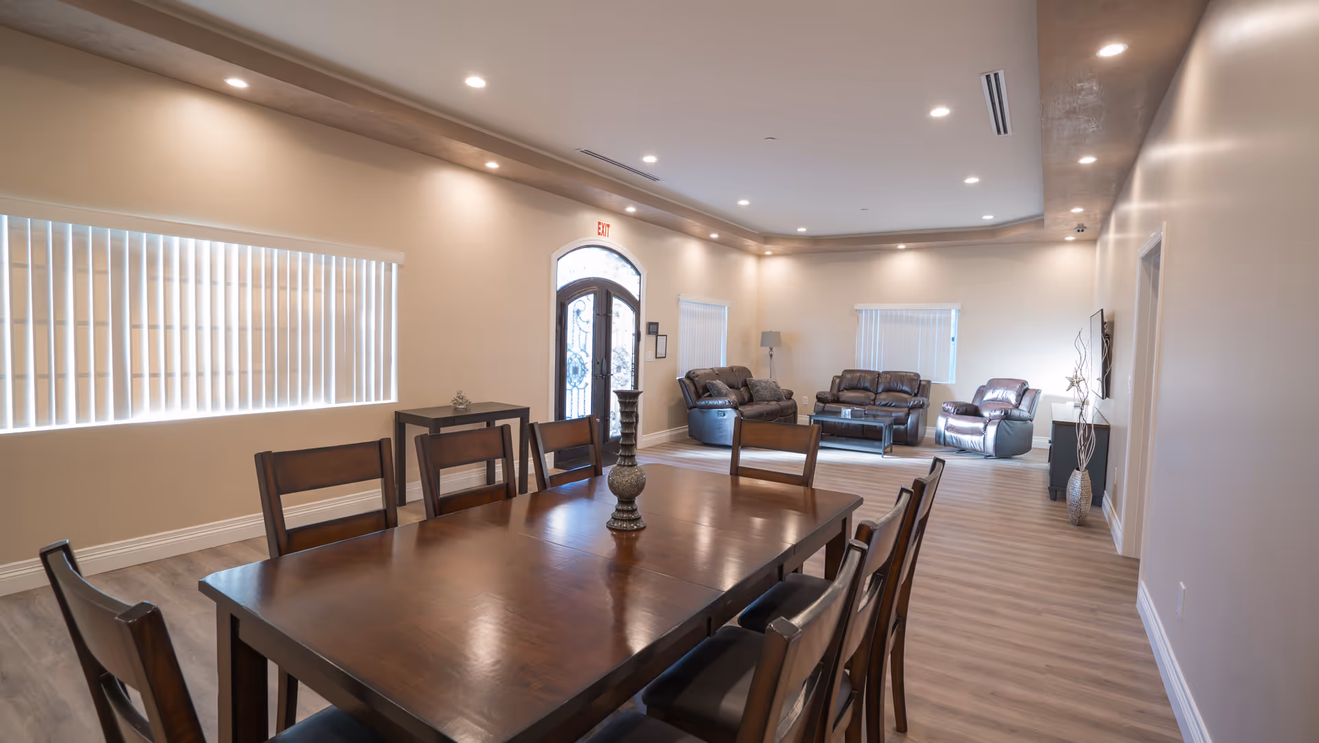 A spacious senior living common area featuring a large wooden dining table with eight chairs in the foreground and a seating area with three leather recliners and a coffee table in the background. The room has light-colored walls, recessed ceiling lights, vertical blinds on the windows, and a decorative glass double door with an exit sign above it.