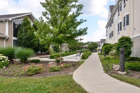 A landscaped outdoor walkway at Cedar Ridge of Fort Wayne, featuring a concrete path bordered by green grass, shrubs, flowers, and trees. Benches and a gazebo are visible along the path, with multi-story residential buildings on the right side under a partly cloudy sky.