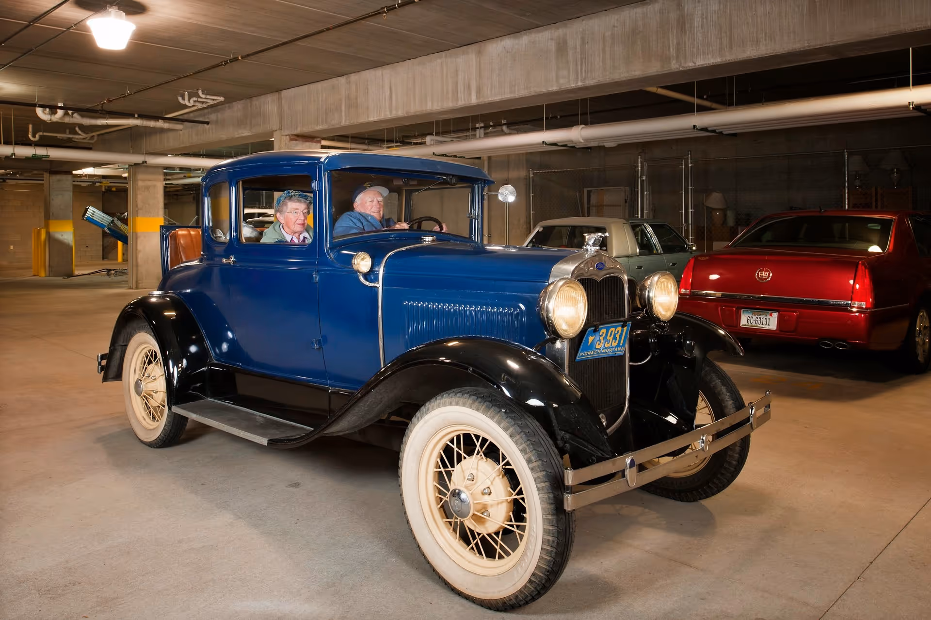 A vintage blue car parked in an underground garage with two elderly people seated inside and modern cars in the background.