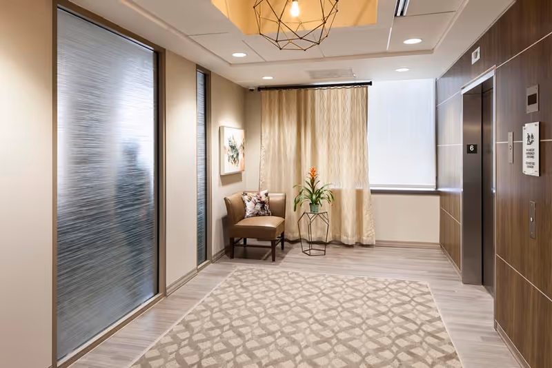 A modern interior hallway area with a patterned carpet, a single brown chair with a decorative pillow, a small table holding a potted plant with orange flowers, beige curtains covering a window, and an elevator with closed doors on the right side. The walls are light-colored with wood paneling around the elevator, and there is a geometric light fixture on the ceiling.