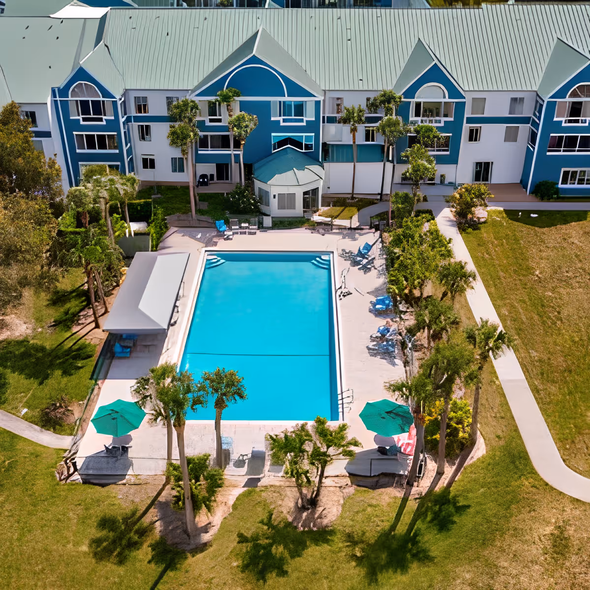 Aerial view of an outdoor swimming pool surrounded by lounge chairs, umbrellas, and palm trees, with a multi-story residential building in the background at Discovery Village Vero Beach.