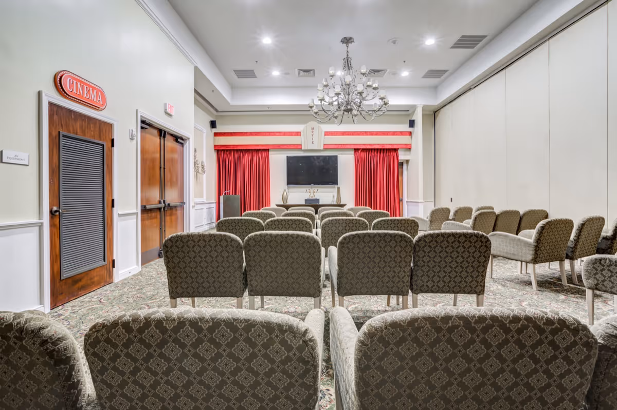Interior view of a small cinema room with patterned upholstered chairs arranged in rows facing a flat screen TV mounted on the wall. The room features red curtains, a chandelier hanging from the ceiling, and a sign above a wooden door labeled 'CINEMA'.