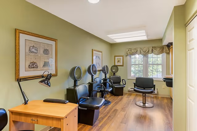 Interior view of a salon area in a senior living facility with three black salon chairs and hair dryers lined up against a green wall. There is a wooden desk with a black lamp on the left side and framed artwork hanging on the wall. A window with a valance lets in natural light at the far end of the room.