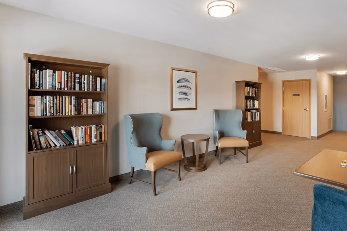 A quiet reading or sitting area in a senior living facility hallway featuring two blue and tan upholstered armchairs with a small round wooden table between them. There are two wooden bookshelves filled with books on either side of the chairs. A framed picture of feathers hangs on the beige wall above the chairs. The carpeted floor and ceiling lights create a warm and inviting atmosphere.