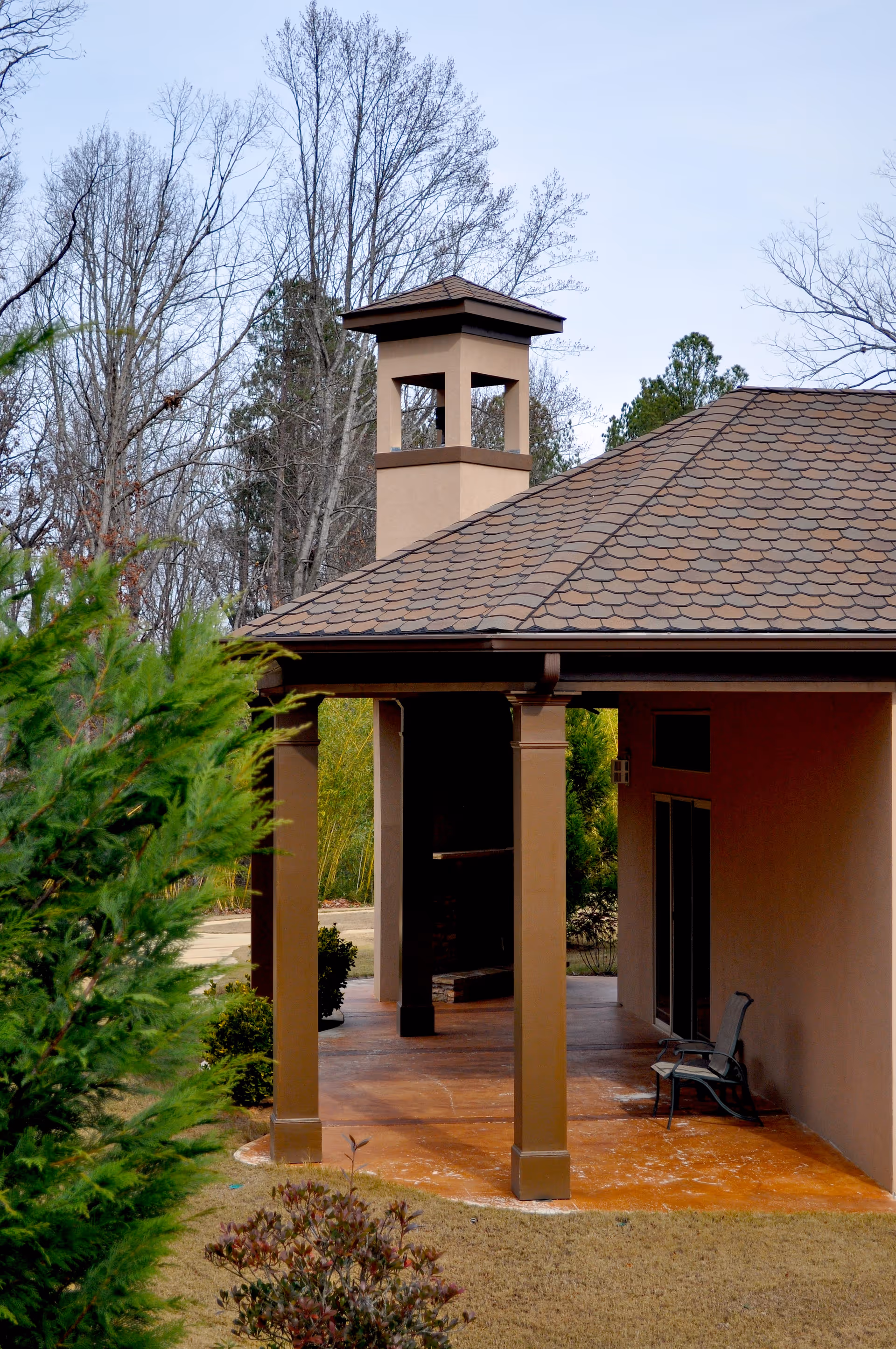 Covered porch entrance of a tan building with columns, a shingled roof and surrounding trees.