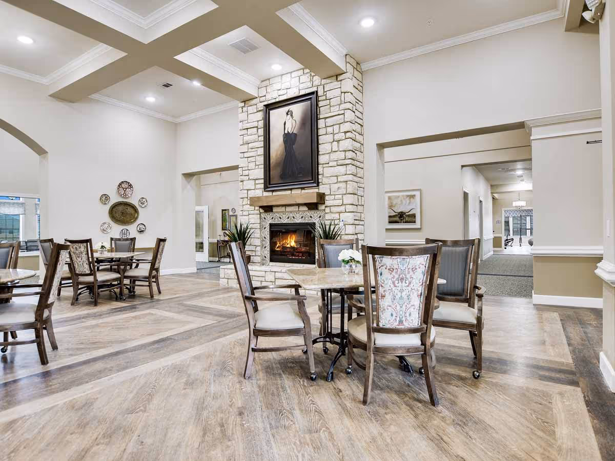 A spacious and well-lit common area with wooden flooring, several round tables with chairs, a stone fireplace with a fire burning, and a framed painting above the fireplace. The walls are light-colored, and there are decorative plates on one wall. The room has a high ceiling with recessed lighting.