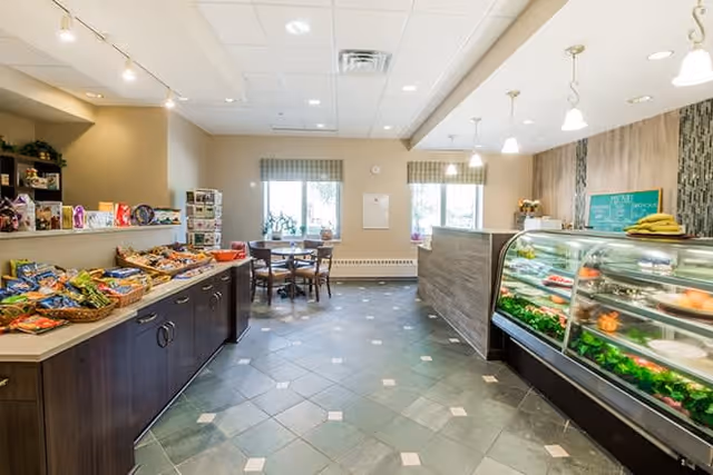 Interior view of a bright and clean dining area in a senior living facility with a display case containing food on the right, a counter with snacks on the left, and a small dining table with chairs near two windows in the background.