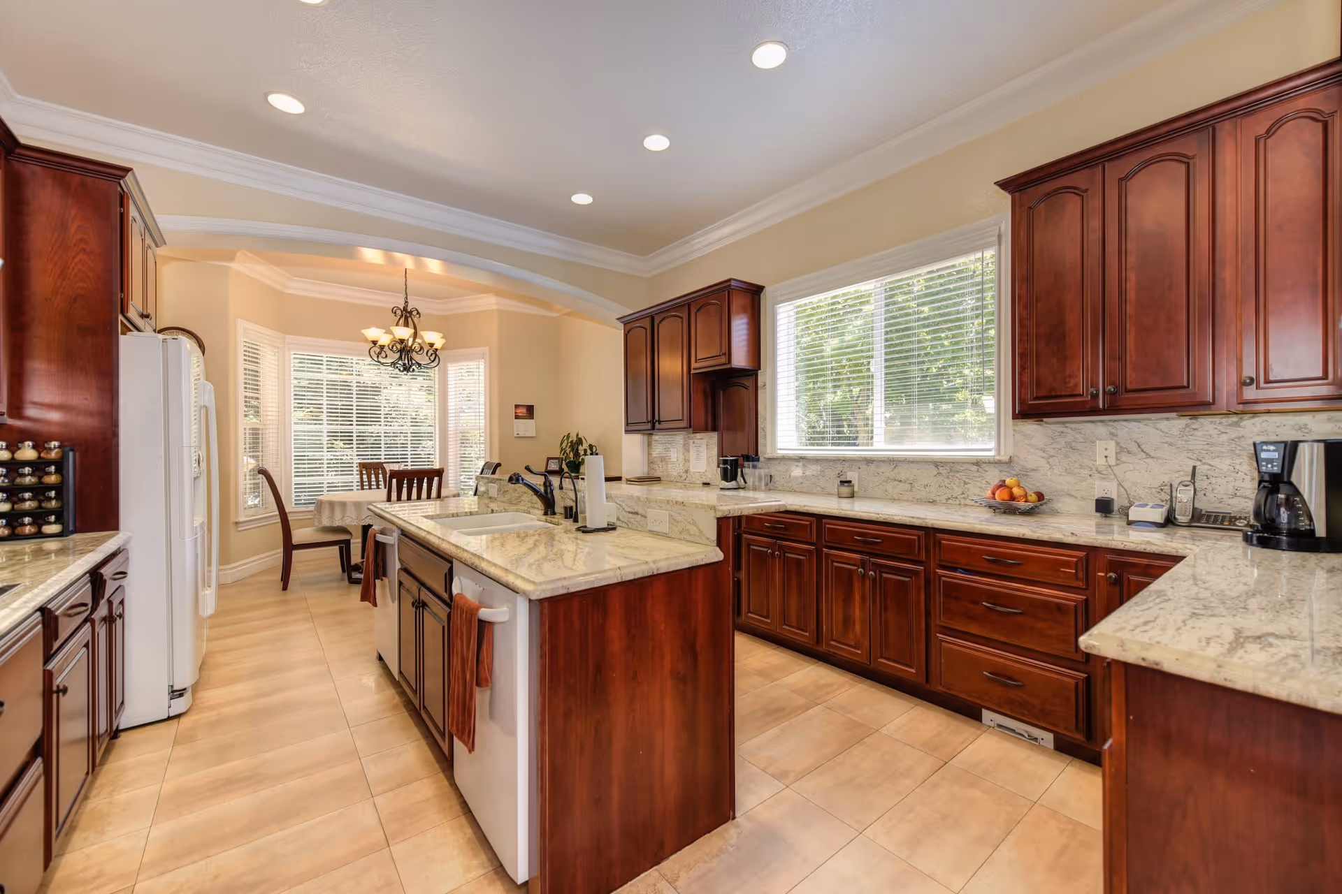 A spacious kitchen with dark wood cabinets, light marble countertops, and a central island with a sink. There is a white refrigerator on the left side and a dining area with a table and chairs near large windows with blinds. The floor is tiled, and the kitchen is well-lit with recessed ceiling lights and a chandelier over the dining table.
