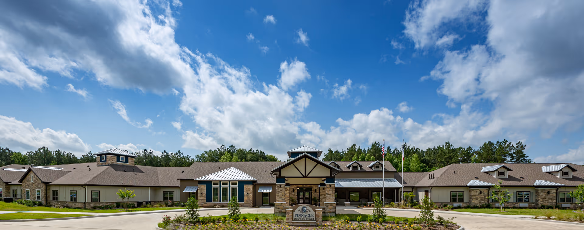 Wide exterior view of Pinnacle Senior Living facility showing a single-story building with a combination of stone and siding facade, multiple windows, and a covered entrance under a partly cloudy blue sky.