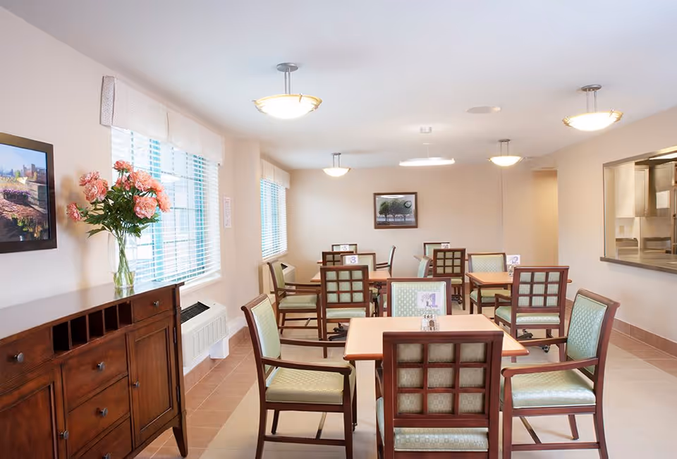 A bright dining room with several wooden tables and chairs with green cushions. There are large windows with white blinds letting in natural light. A wooden sideboard with a vase of pink flowers is on the left, and a framed picture hangs on the wall. The room has ceiling lights and a serving window to the kitchen on the right.