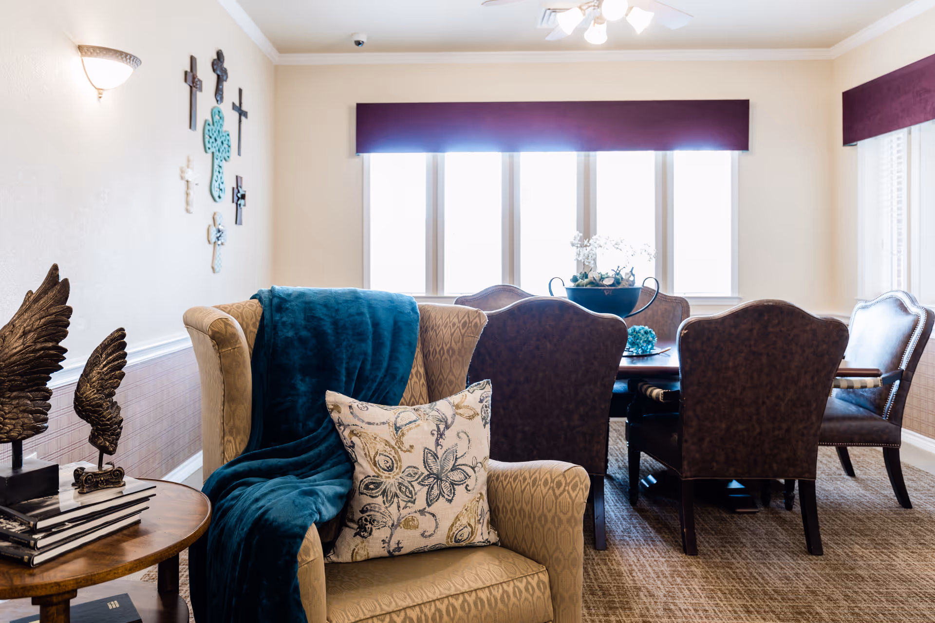 A cozy and well-lit living room area with a beige armchair draped with a teal throw blanket and a decorative pillow. Behind the armchair is a wooden table with two wing sculptures and stacked books. In the background, there is a dining table surrounded by six dark upholstered chairs, with a decorative centerpiece on the table. The room has large windows with purple valances and a wall decorated with various crosses.
