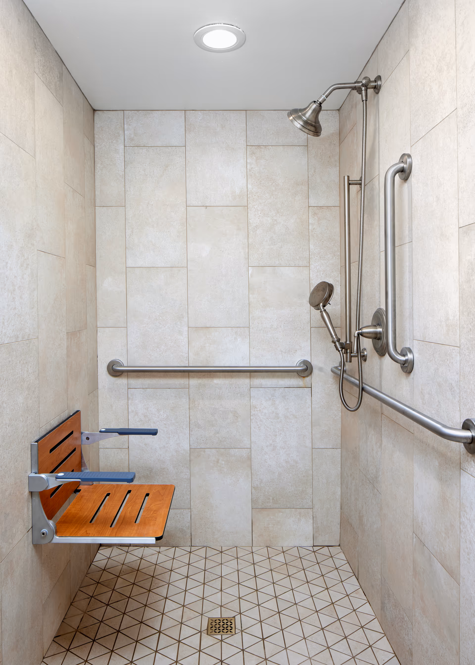 Tiled accessible shower with a fold-down wooden seat, multiple grab bars, and overhead and handheld showerheads.