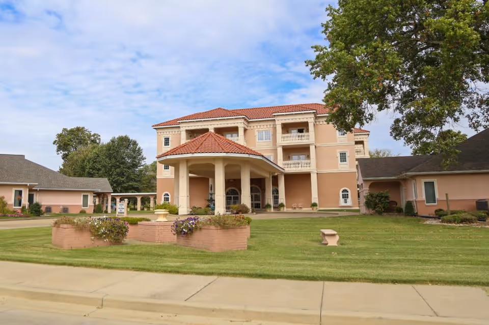 Exterior view of Fern Terra of Owensboro, a multi-story senior living facility with a covered entrance, beige and peach colored walls, red tiled roof, surrounded by green lawn, flower beds, trees, and a clear sky with some clouds.