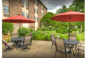 Outdoor patio courtyard with round tables, chairs, and red umbrellas beside a multi-story brick building and landscaped greenery.