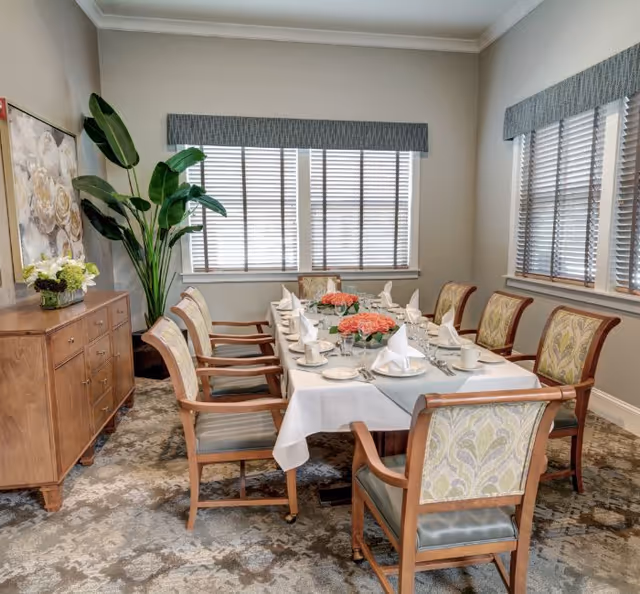 Formal dining room with a table set for eight, floral centerpieces, wooden chairs, a sideboard, and large windows with blinds.