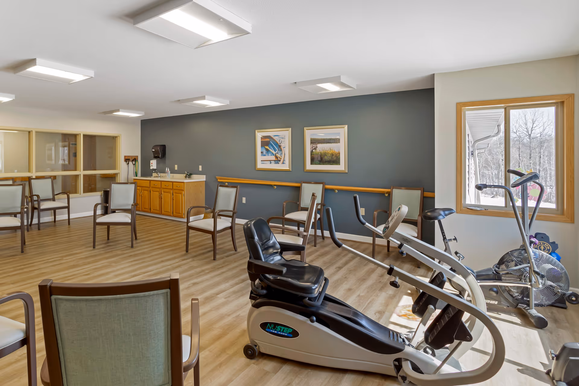 A bright senior living activity room with exercise equipment and arranged chairs, a small kitchenette, framed artwork on the wall, and a window showing snowy trees.