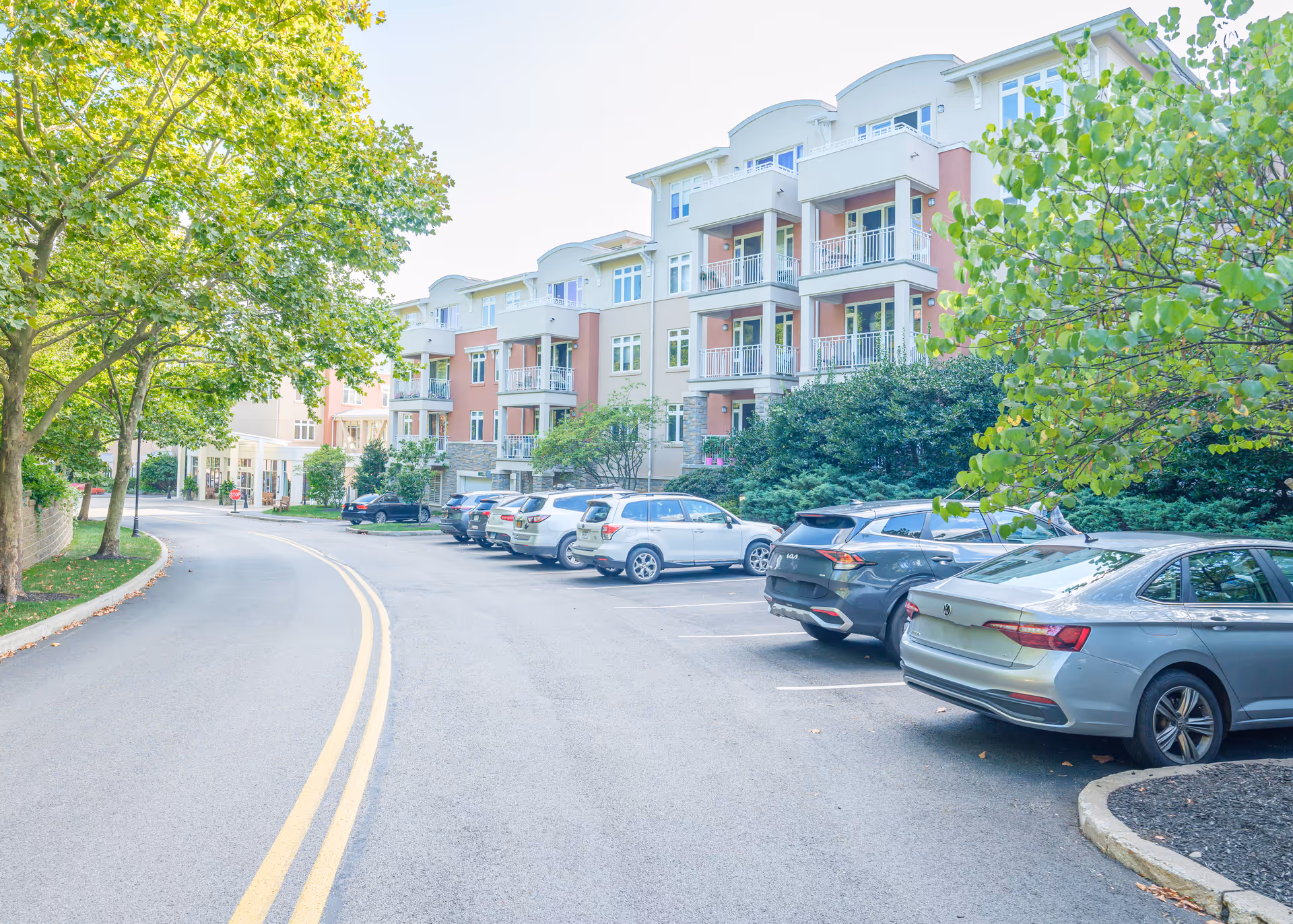 Exterior view of Kendal On Hudson senior living facility showing a multi-story building with balconies, a parking lot with several parked cars, and a tree-lined road with double yellow lines.