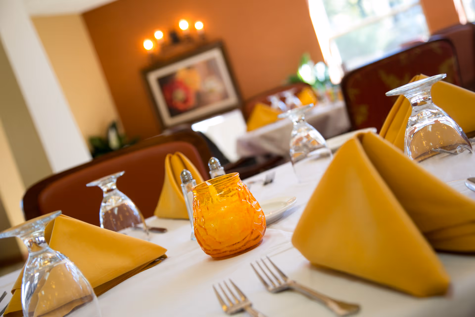 Close-up view of a dining table set with yellow folded napkins, upside-down wine glasses, silver forks, and an orange glass candle holder in a warmly lit dining room with brown chairs and a framed picture on the wall.