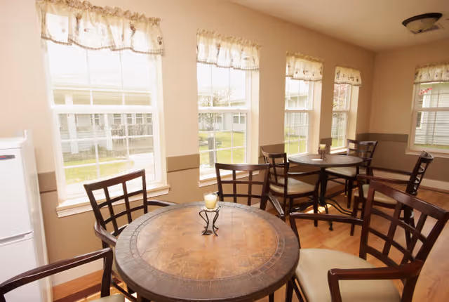 A bright dining area with round wooden tables and cushioned chairs arranged near large windows with floral valances, allowing natural light to fill the room. A small candle holder is placed on the closest table.