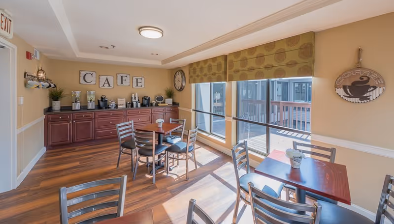 Sunlit café-style dining room with wooden tables and chairs, a countertop with coffee machines and cabinets under a 'CAFE' sign, and large windows.