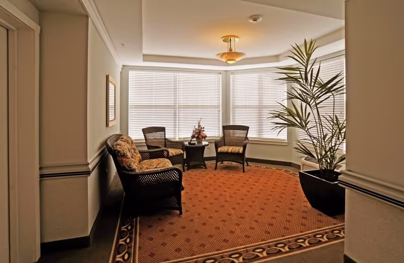A cozy sitting area with three wicker chairs and a wicker loveseat arranged around a small round table with a flower arrangement. The space has large windows with blinds, a patterned carpet, and a tall potted plant in the corner.