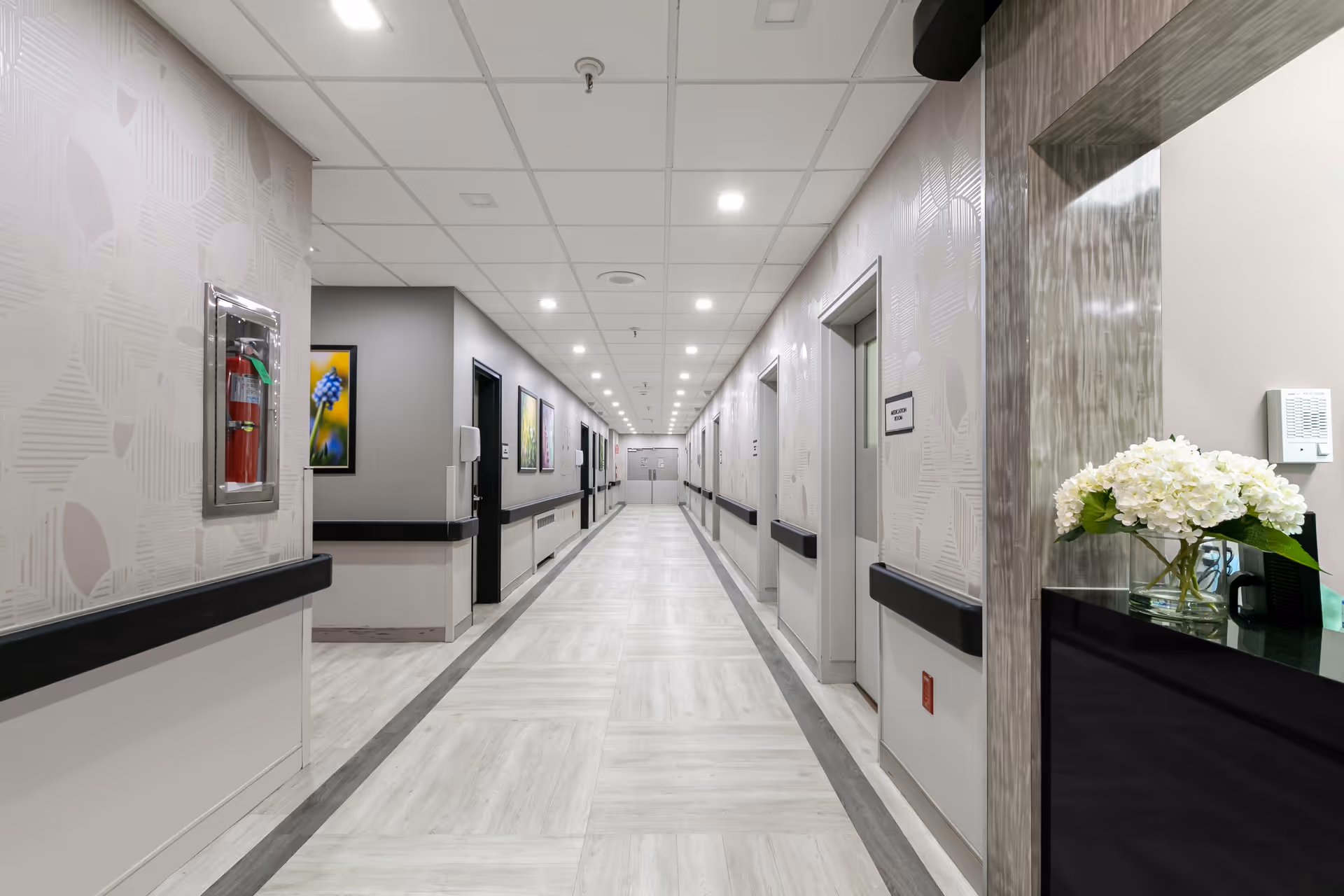 A long, well-lit hallway in a senior living facility with light-colored flooring and walls. The hallway has multiple doors on both sides, handrails along the walls, framed artwork, and a fire extinguisher mounted on the left wall. On the right side near the foreground, there is a counter with a vase of white flowers.