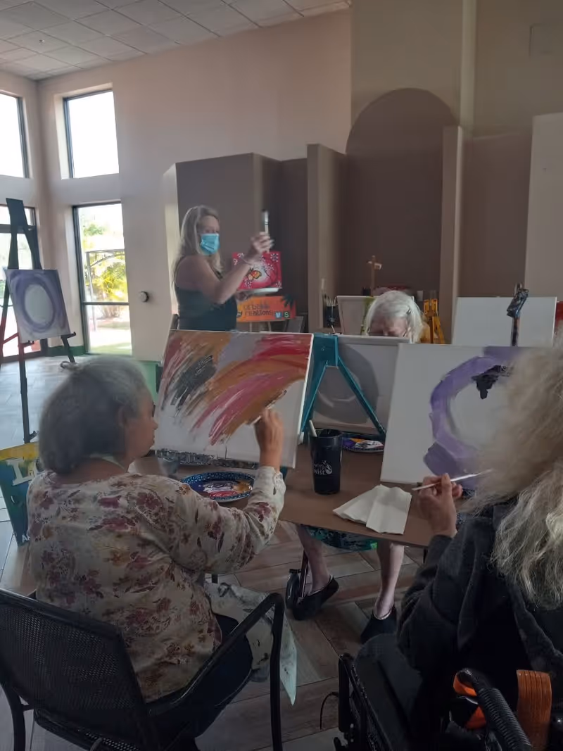 A group of elderly women seated around a table engaged in painting on canvases in a well-lit room with large windows. One woman is standing and wearing a face mask, holding up a painting. The atmosphere appears to be a creative art session.