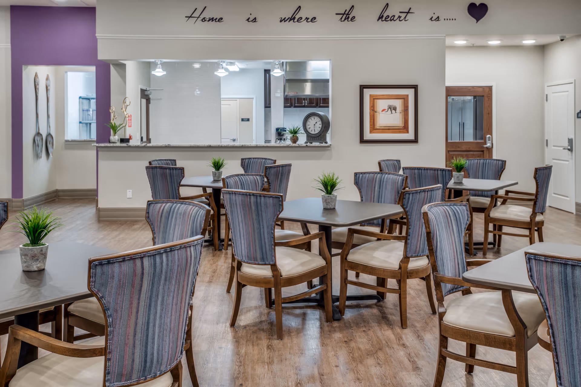 A dining area in a senior living facility with several tables and chairs arranged neatly. Each table has a small potted plant as decoration. The room has wood flooring and a serving window to the kitchen area. On the wall above the serving window is a decorative phrase that reads 'Home is where the heart is...' with a heart symbol. There is framed artwork on the wall and a door leading to another room.