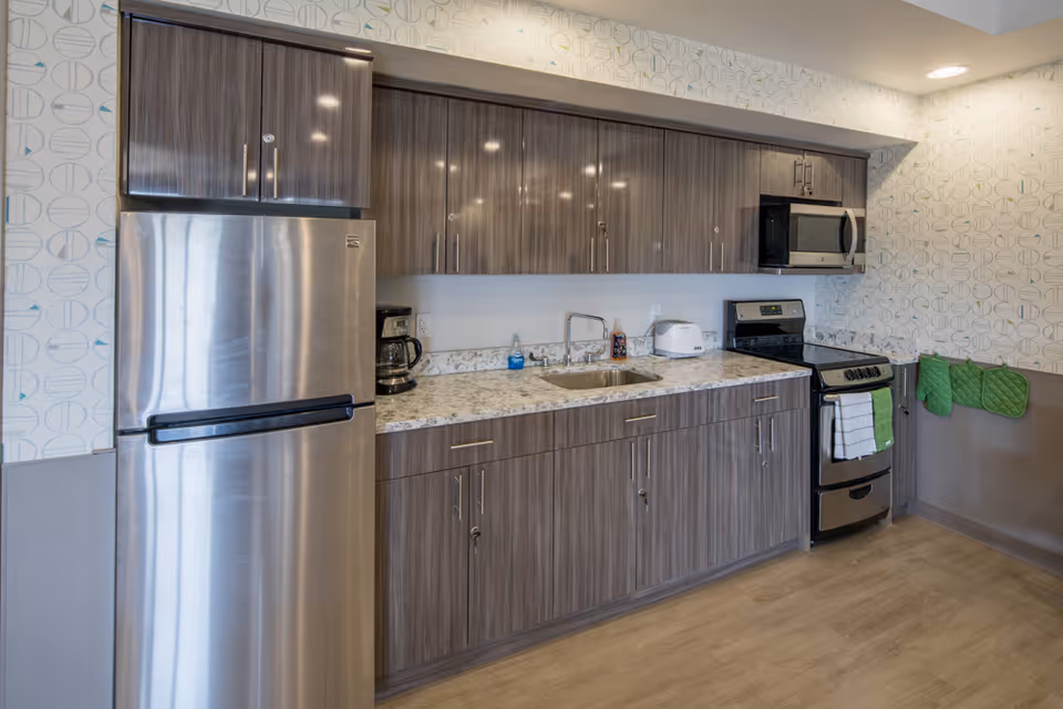 Modern kitchen area with stainless steel refrigerator, stove, and microwave. The kitchen has wood-grain cabinets, a granite countertop with a sink, a coffee maker, toaster, and green oven mitts hanging on the wall. The walls have a patterned wallpaper and the floor is light wood.