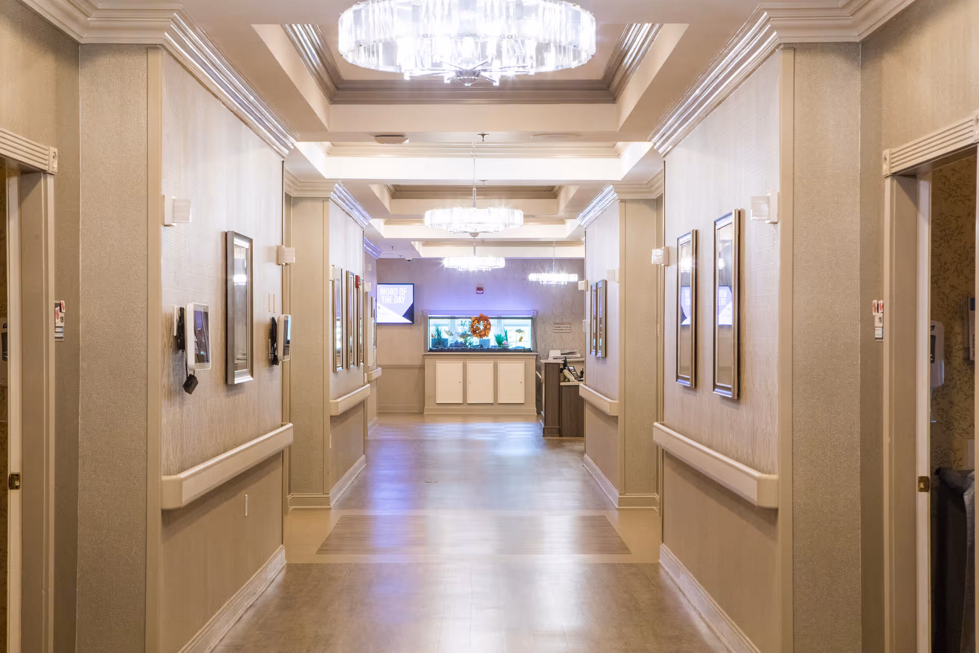 A well-lit hallway in a senior living facility with beige walls, framed pictures on the walls, handrails on both sides, and multiple ceiling lights. At the end of the hallway, there is a fish tank and a small reception or desk area.