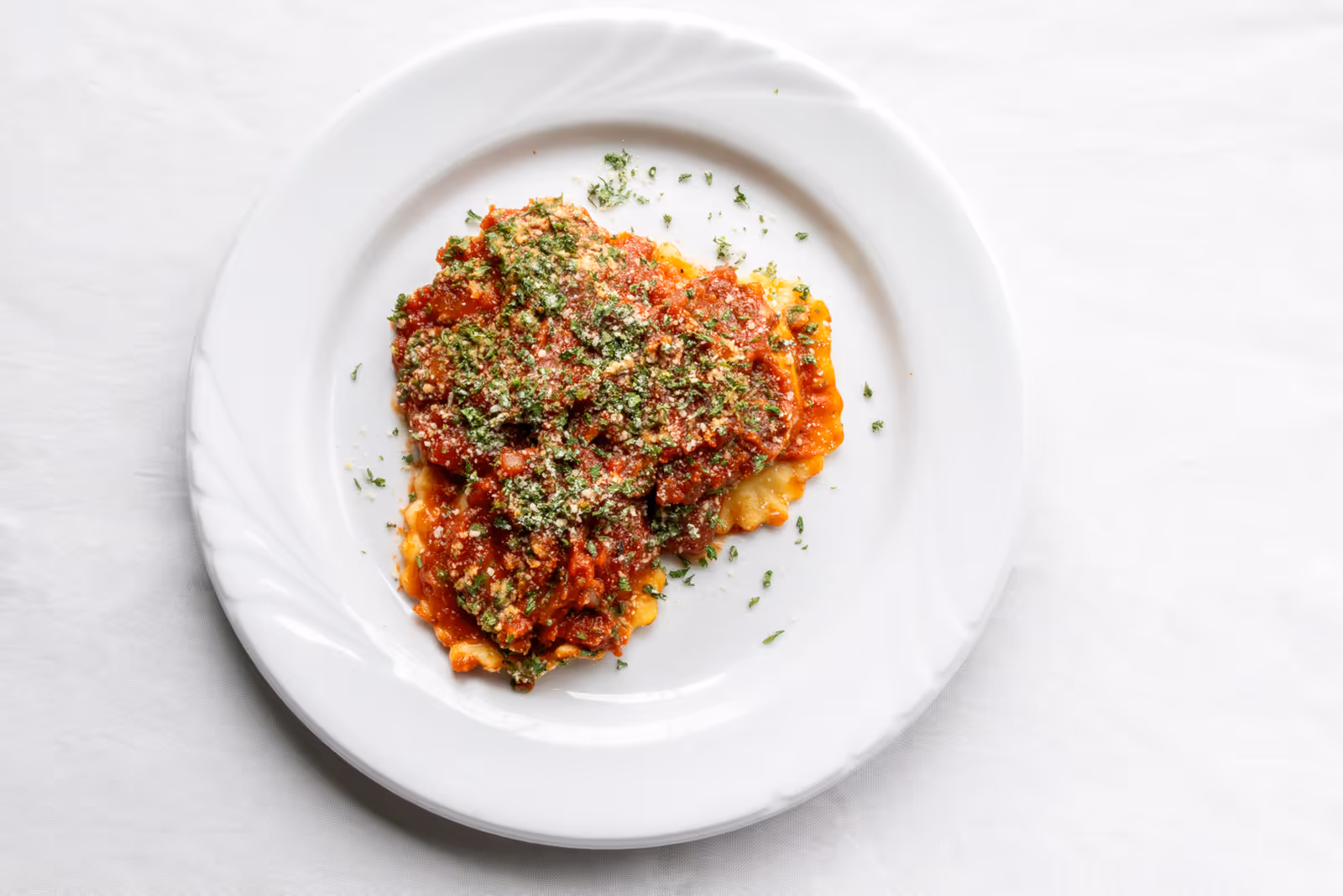 White plate with ravioli-like pasta topped with tomato sauce, grated cheese, and chopped herbs on a white tablecloth.