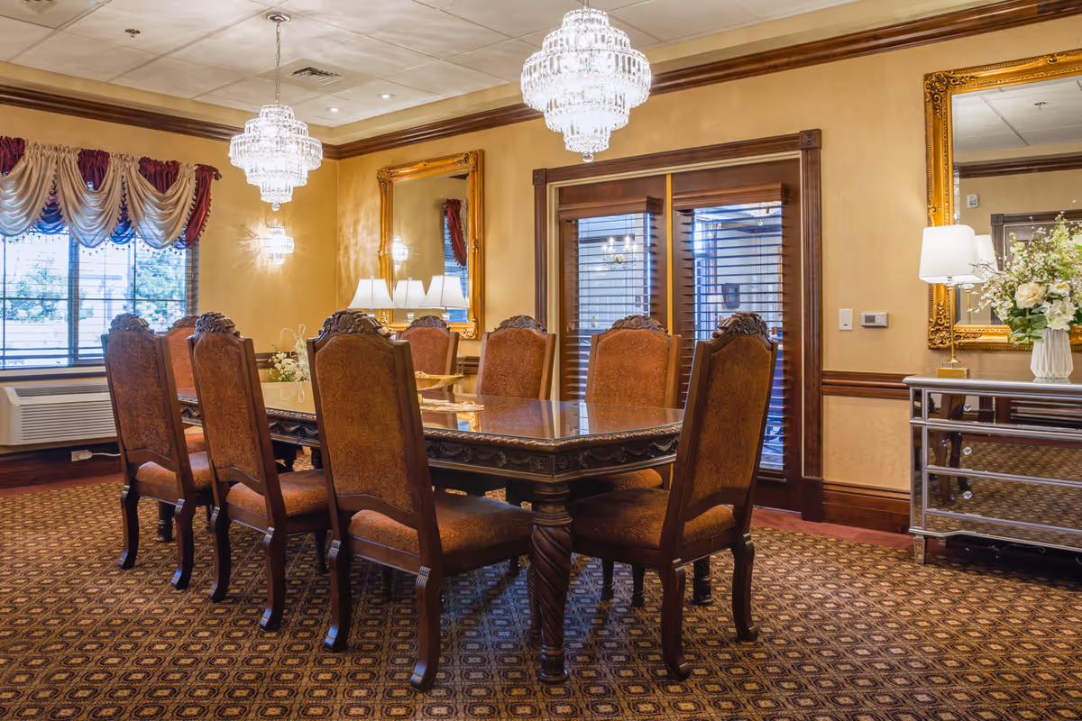 A formal dining room with a large wooden dining table surrounded by eight upholstered chairs. The room features ornate chandeliers hanging from the ceiling, large mirrors with gold frames on the walls, a window with decorative curtains, and a sideboard with a lamp and floral arrangement.