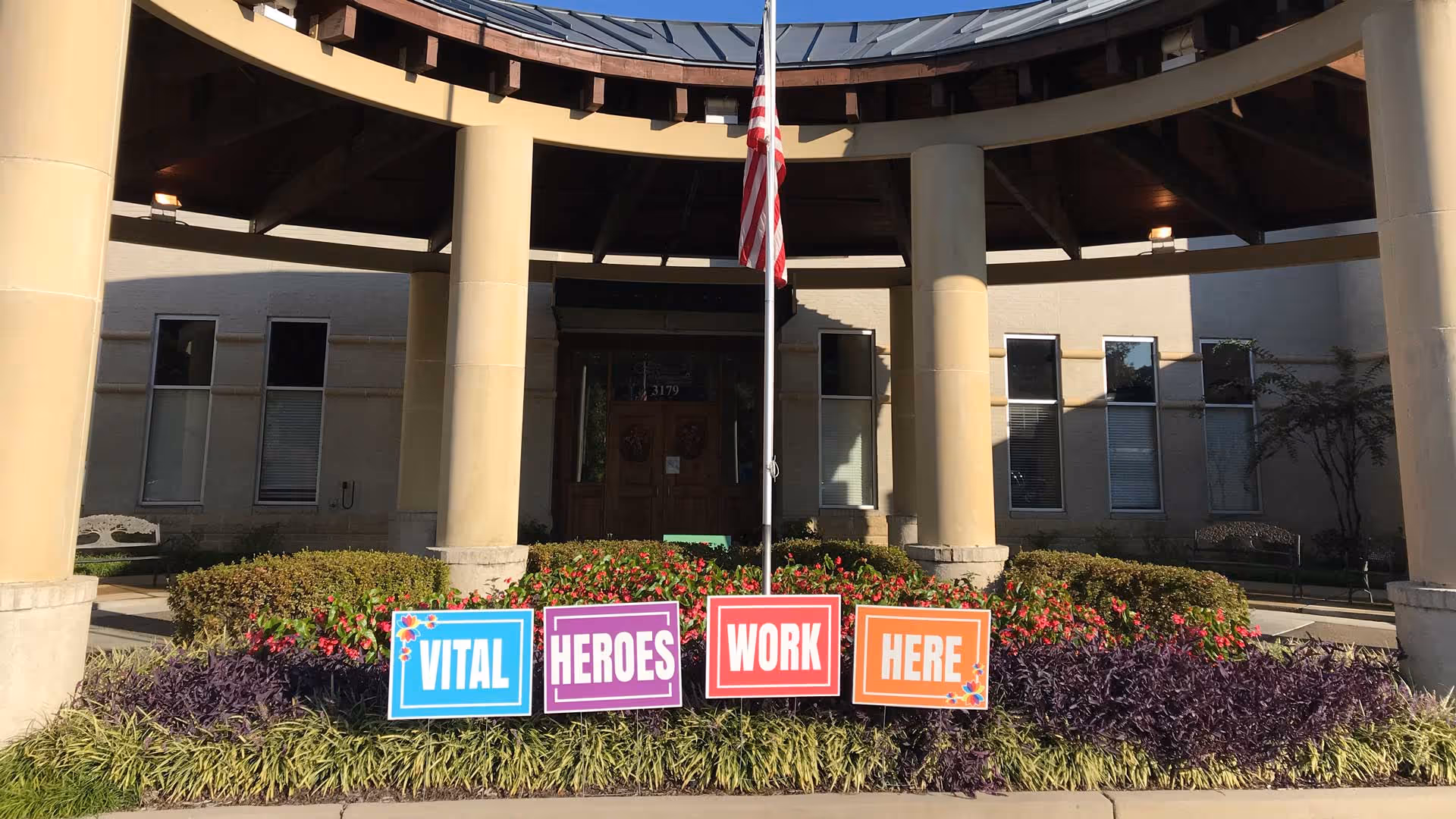 Entrance of a building with large beige columns supporting a circular roof. In front of the entrance, there is a flagpole with an American flag. Below the flagpole, colorful signs placed in the garden read 'VITAL HEROES WORK HERE'. The building has several windows and a double wooden door.