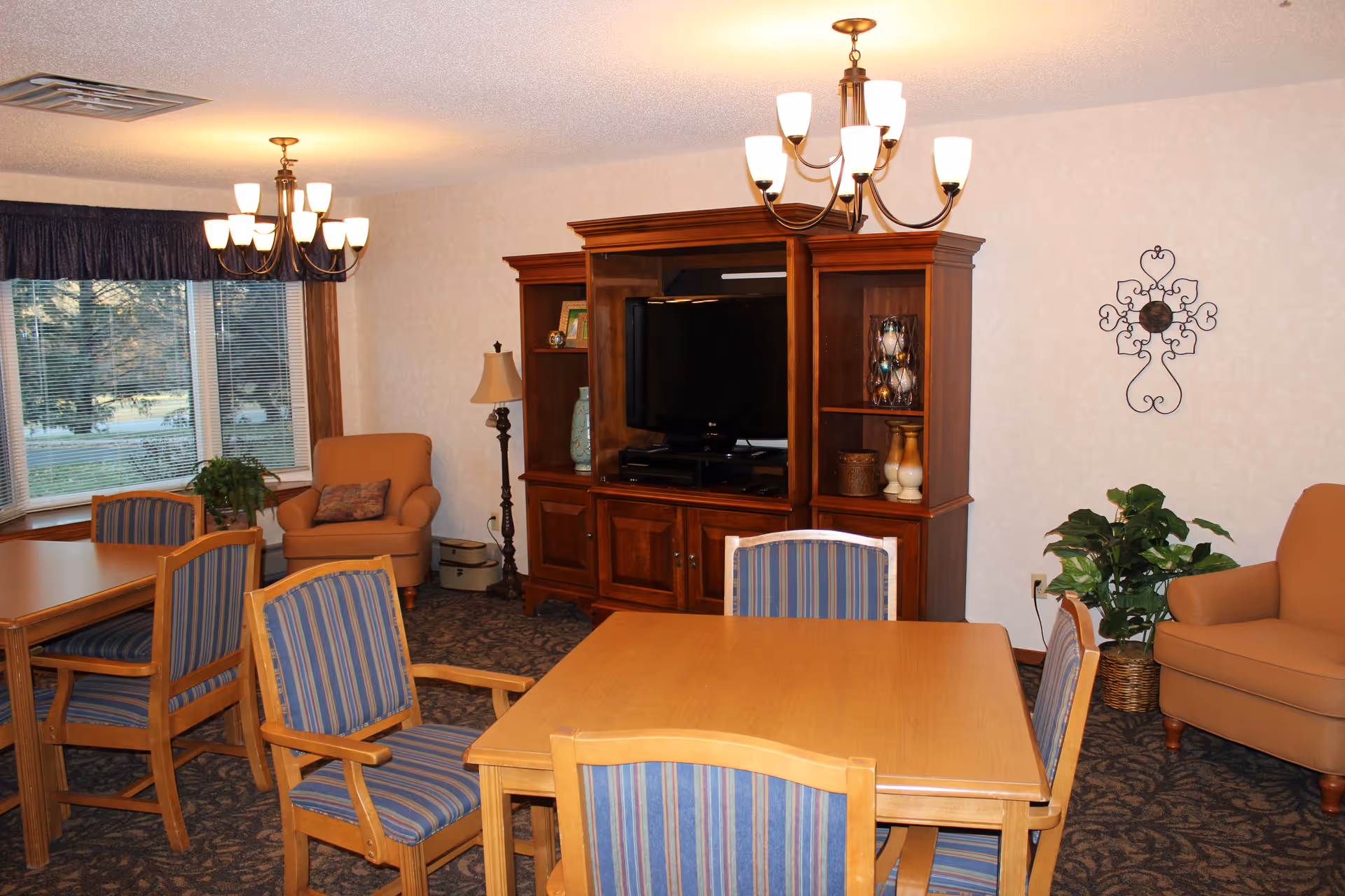 Communal dining and sitting area with wooden tables and striped chairs, armchairs, and a large entertainment cabinet with a TV.