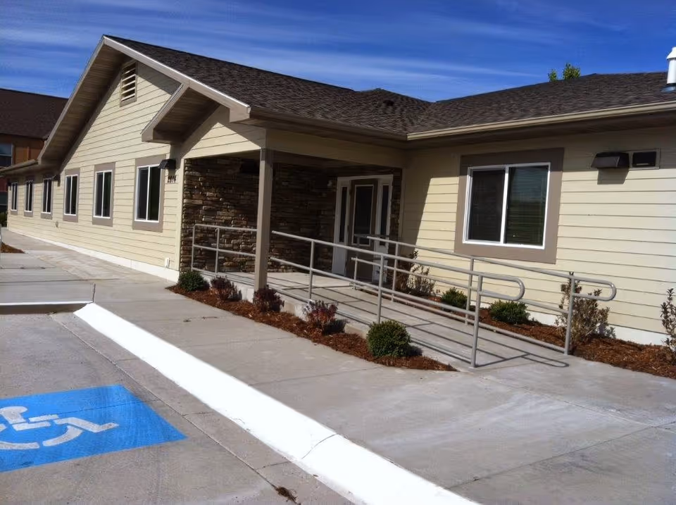 Front exterior of a single-story assisted living building showing the entrance with an accessibility ramp and a marked handicapped parking space.