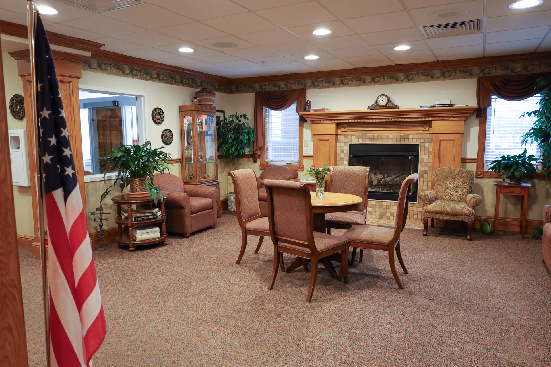 A cozy living room area in The Winslow Independent Living facility featuring a round wooden table with four upholstered chairs, a fireplace with a wooden mantle, two windows with brown curtains, several armchairs, potted plants, and an American flag in the foreground.
