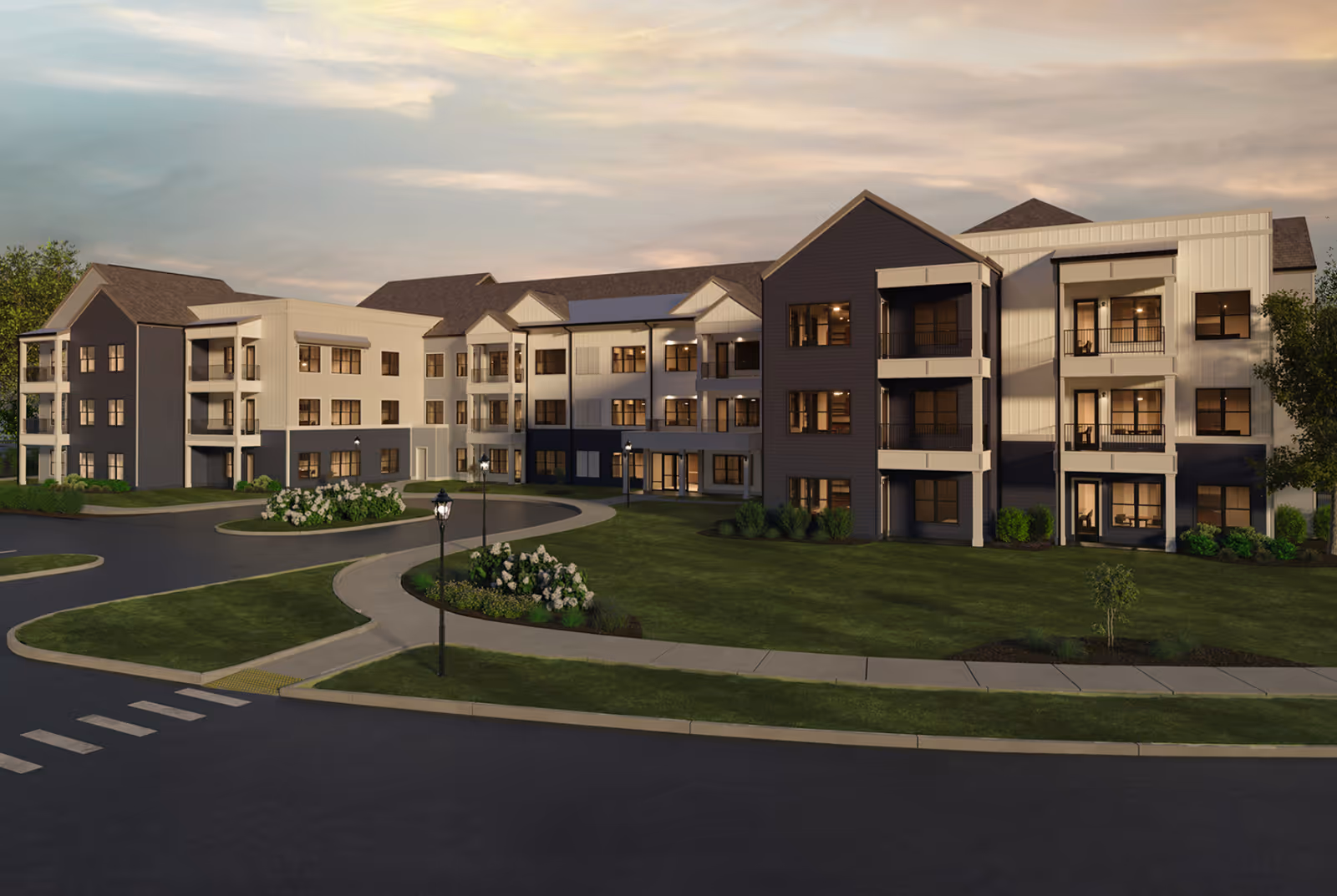 Exterior view of a modern three-story senior living facility building with multiple windows and balconies, surrounded by well-maintained green lawns, flower beds, and a curved driveway with street lamps under a partly cloudy sky at dusk.